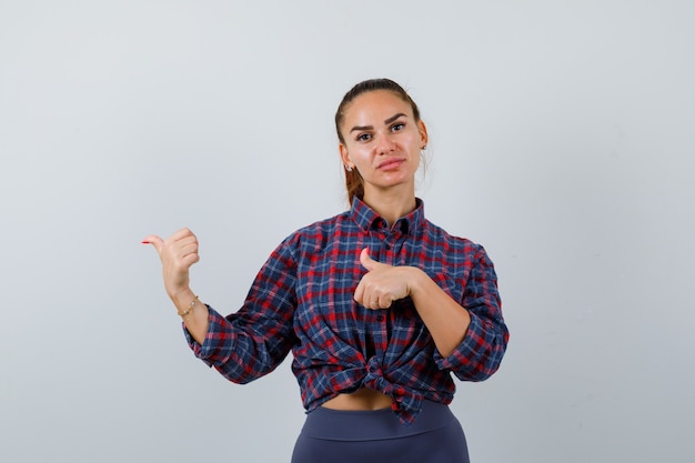 Jonge vrouw die duimen naar de linkerkant wijst in een geruit hemd, broek en er zelfverzekerd uitziet, vooraanzicht.