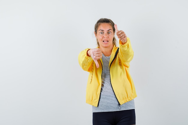 Jonge vrouw die duimen op en neer in t-shirt, jasje toont en verbaasd, vooraanzicht kijkt.