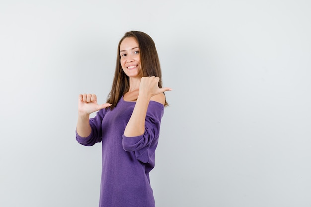 Jonge vrouw die duimen terug in violet overhemd richt en gelukkig kijkt. vooraanzicht.