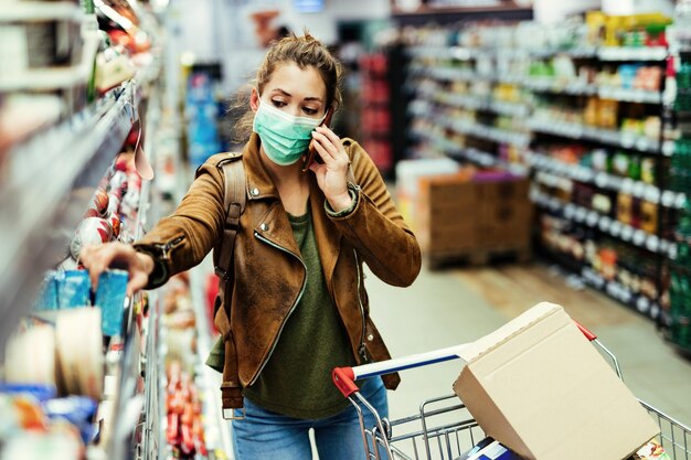 Jonge vrouw die een beschermend gezichtsmasker draagt terwijl ze boodschappen doet en telefoneert tijdens de coronaviruspandemie in de supermarkt