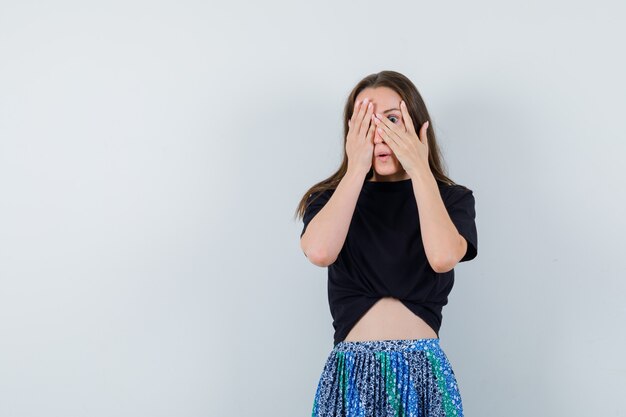 Jonge vrouw die een deel van het gezicht bedekt met handen en door vingers in zwart t-shirt en blauwe rok kijkt en aantrekkelijk kijkt