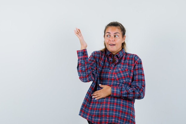 Jonge vrouw die een hand op de buik rust terwijl ze een andere hand uitrekt en ernaar kijkt in een geruit overhemd en er mooi uitziet