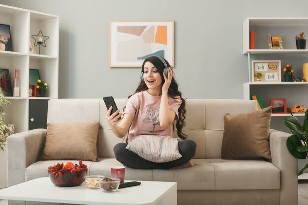 Jonge vrouw die een koptelefoon draagt en naar een telefoon kijkt die op de bank zit achter de salontafel in de woonkamer