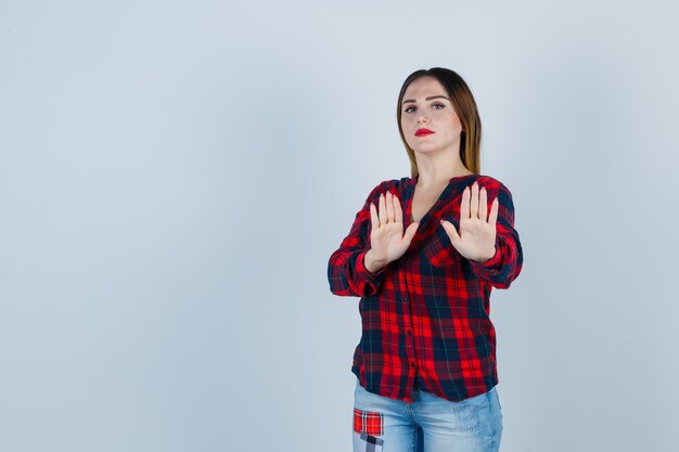 Jonge vrouw die een stopgebaar in een geruit overhemd toont en er terughoudend uitziet. vooraanzicht.