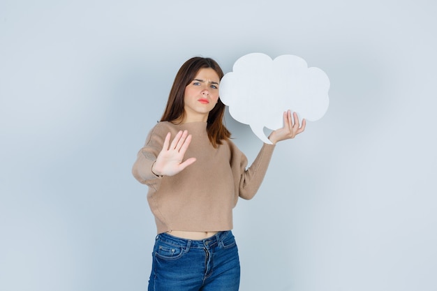 Jonge vrouw die een stopgebaar toont, een papieren tekstballon in een trui, jeans houdt en er angstig uitziet, vooraanzicht.