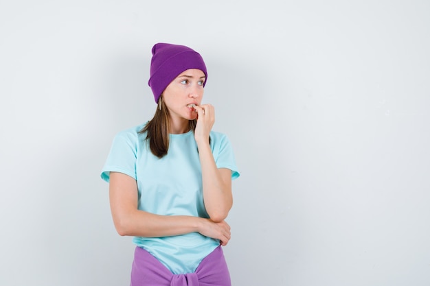 Jonge vrouw die emotioneel vingers bijt in blauw t-shirt, paarse muts en er angstig uitziet. vooraanzicht.