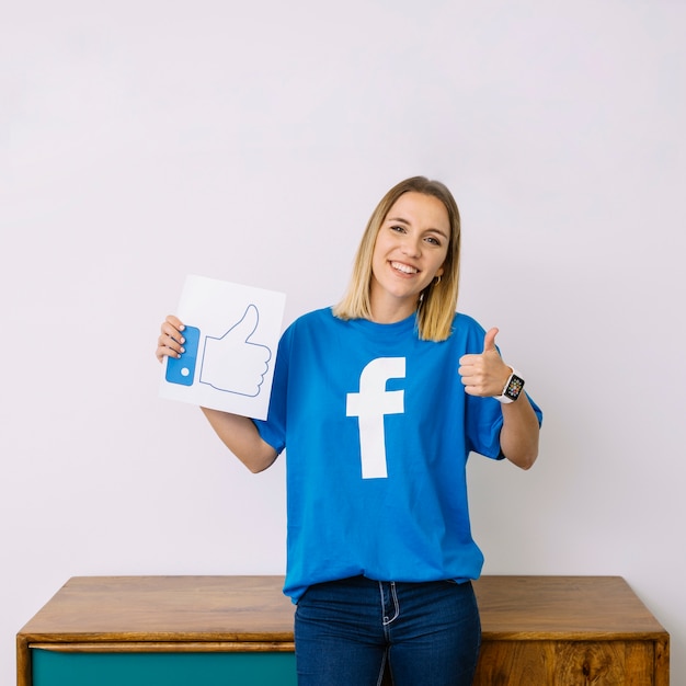 Jonge vrouw die facebook-t-shirtholding zoals pictogram draagt ​​dat thumbup teken toont