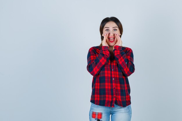 Jonge vrouw die geheim vertelt terwijl ze haar handen in de buurt van de mond houdt in een geruit overhemd, een spijkerbroek en er opgewonden uitziet. vooraanzicht.