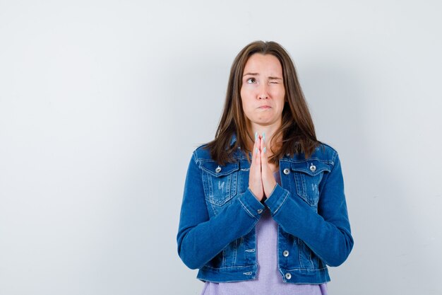 Jonge vrouw die geklemde handen toont in een smekend gebaar, omhoog kijkt in een spijkerjasje en er hoopvol uitziet. vooraanzicht.