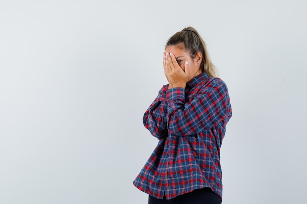 Jonge vrouw die gezicht bedekt met handen, door vingers in geruit overhemd kijkt en bang kijkt