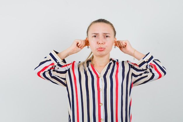 Jonge vrouw die haar handen aan de oren drukt en haar lippen in gestreepte blouse buigt en verontrust kijkt