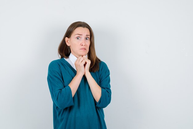 Jonge vrouw die haar handen onder de kin in een trui over een wit overhemd houdt en er bang uitziet, vooraanzicht.