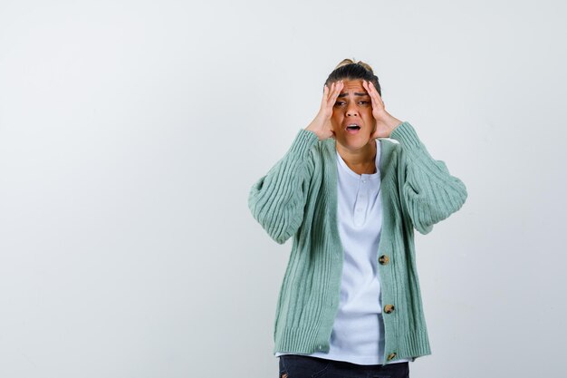 Jonge vrouw die haar handen onder ogen ziet, grimassen trekt in wit t-shirt en mintgroen vest en er geïrriteerd uitziet