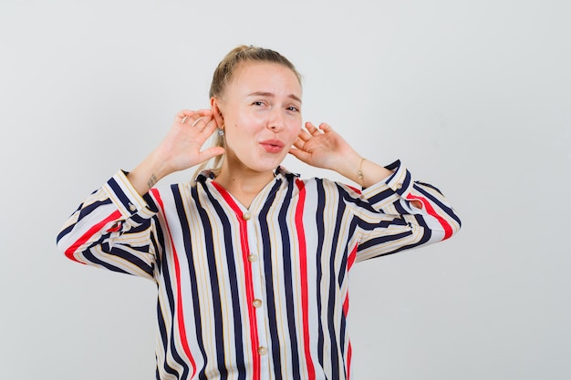 Jonge vrouw die haar handen op de oren in gestreepte blouse drukt en gefrustreerd kijkt