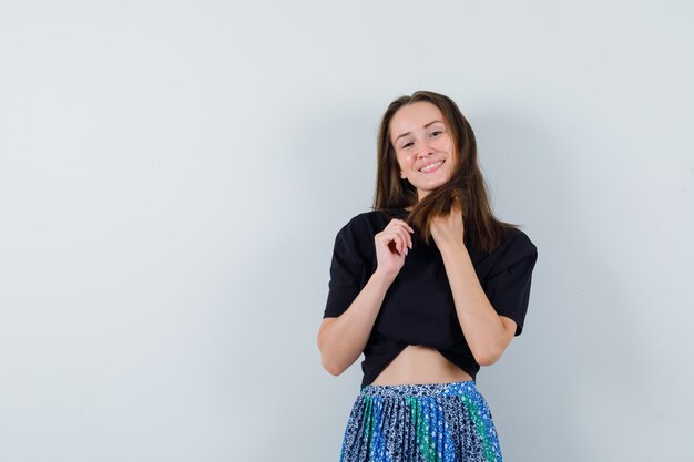 Jonge vrouw die haar met handen instopt, die zich voordeed op camera in zwart t-shirt en blauwe rok en er aantrekkelijk uitziet