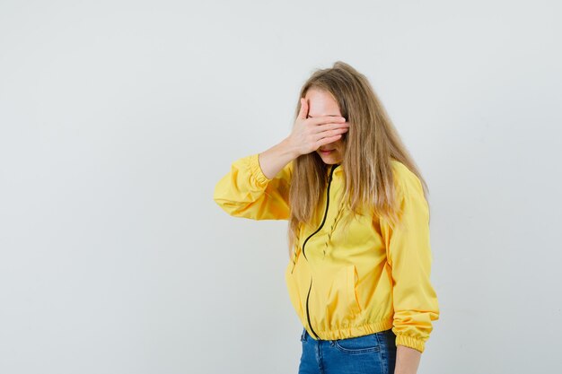 Jonge vrouw die haar ogen bedekt met dient geel bomberjack en blauwe jean in en kijkt verlegen, vooraanzicht.