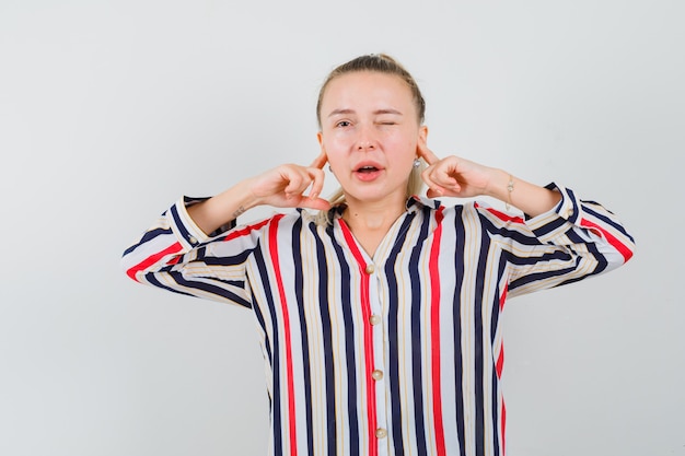 Jonge vrouw die haar oren met haar wijsvingers in gestreepte blouse sluit en geïrriteerd kijkt