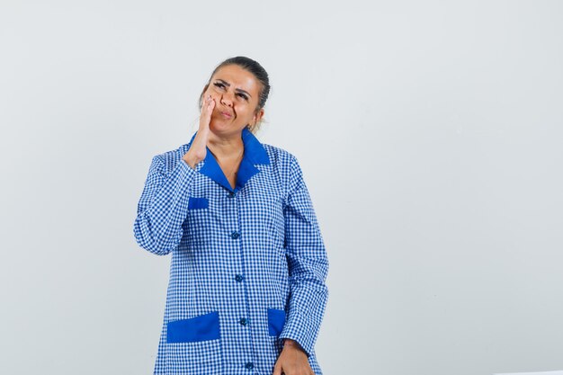 Jonge vrouw die hand dichtbij mond zet, die zich in het denken gebaar in het blauwe overhemd van de gingangpyjama bevindt en peinzend kijkt. vooraanzicht.