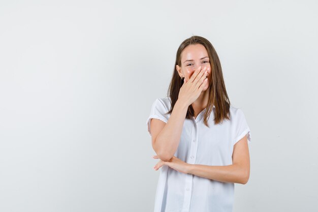 Jonge vrouw die hand op mond behandelt terwijl stiekem in witte blouse lacht en vrolijk kijkt.