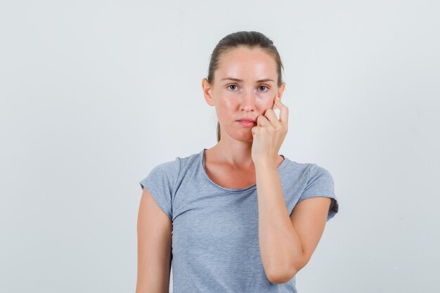 Jonge vrouw die hand op wang in grijs t-shirt houdt en teleurgesteld kijkt. vooraanzicht.