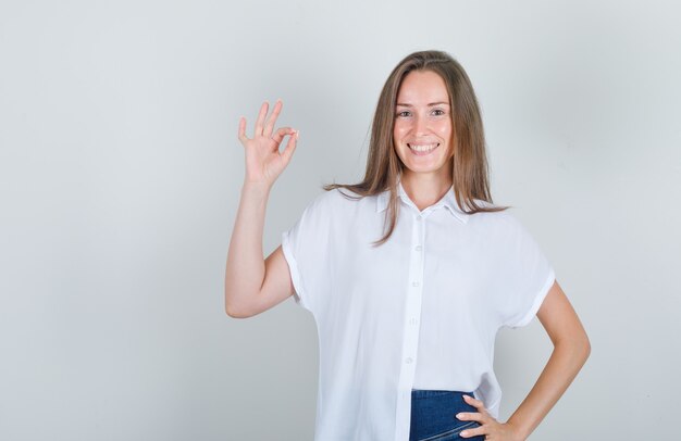 Jonge vrouw die hand opheft en in wit t-shirt glimlacht