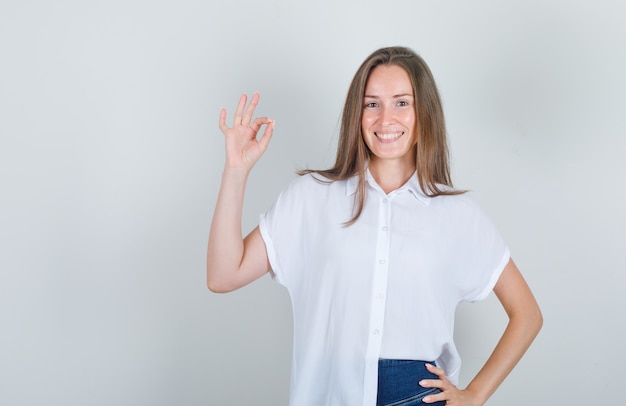 Jonge vrouw die hand opheft en in wit t-shirt glimlacht