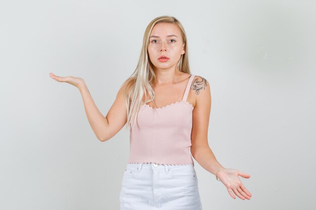 Jonge vrouw die handen met open handpalmen in singlet, minirok houdt en hulpeloos kijkt.