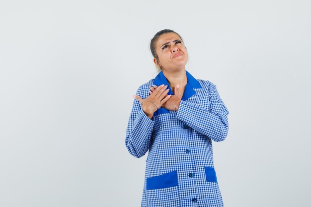 Jonge vrouw die handen op borst rust, die hierboven in blauw geruit pyjamaoverhemd kijkt en peinzend kijkt. vooraanzicht.