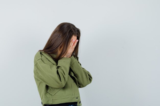 Jonge vrouw die handen op haar gezicht in groene jas behandelt en droevig, vooraanzicht kijkt. ruimte voor tekst