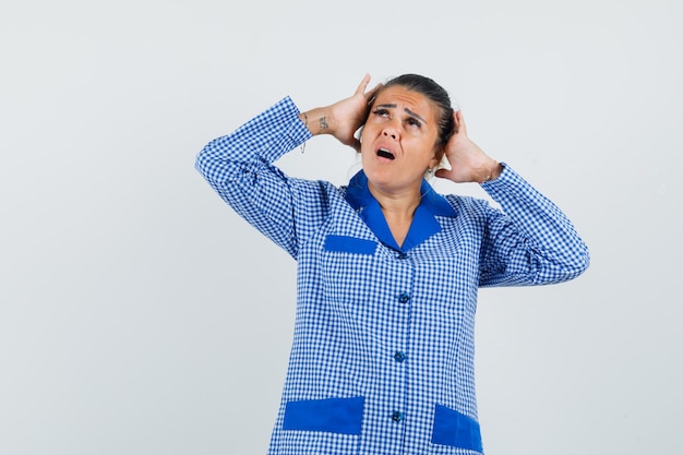 Jonge vrouw die handen op het hoofd zet, hierboven in blauw geruit pyjamaoverhemd kijkt en verbaasd kijkt. vooraanzicht.