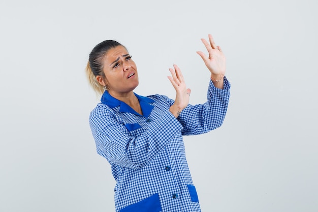 Jonge vrouw die handen opheft als iets in het blauwe overhemd van de gingangpyjama en ziet er moe tegen te houden. vooraanzicht.