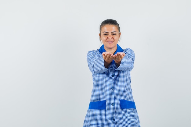 Jonge vrouw die handen uitrekt als uitnodigend om in blauw geruit pyjamaoverhemd te komen en er beminnelijk uitziet. vooraanzicht.