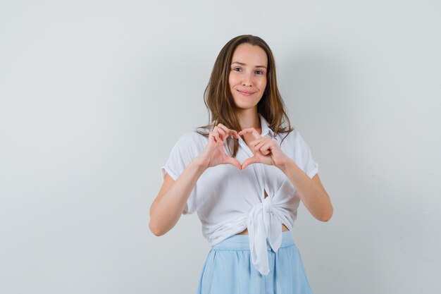 Jonge vrouw die hartvorm met vingers in witte blouse en lichtblauwe rok toont en gelukkig kijkt