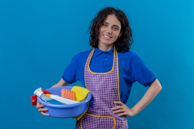 Jonge vrouw die het bassin van de schortholding met schoonmakende hulpmiddelen dragen die het zekere glimlachen vrolijk over blauwe muur kijken