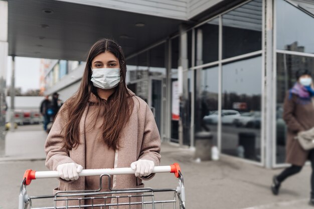 Jonge vrouw die het masker van het beschermingsgezicht draagt tegen coronavirus dat een boodschappenwagentje duwt.