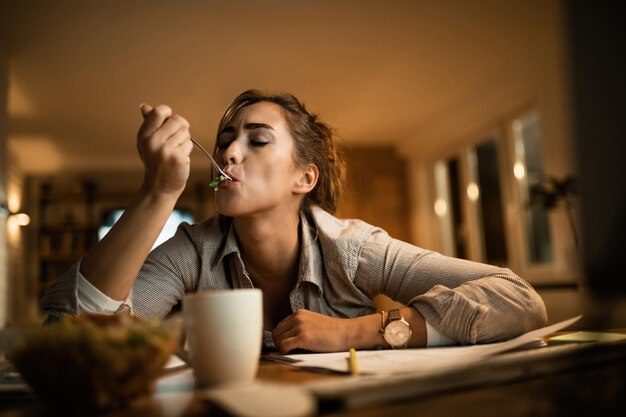 Jonge vrouw die honger heeft en salade eet terwijl ze 's avonds thuis studeert