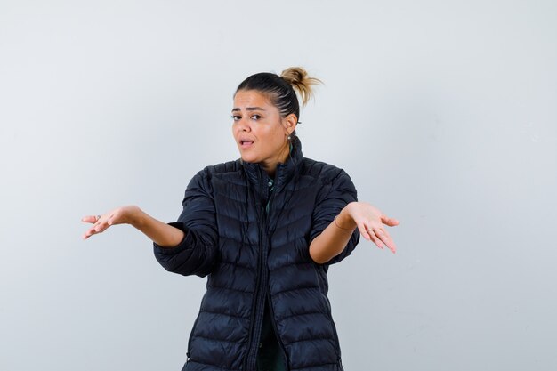 Jonge vrouw die hulpeloos gebaar in pufferjas toont en verward, vooraanzicht kijkt.