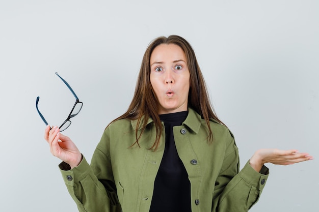 Jonge vrouw die hulpeloos gebaar toont terwijl glazen in groen jasje wordt gehouden en verbaasd, vooraanzicht kijkt.