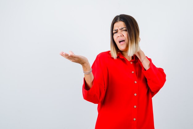 Jonge vrouw die iets laat zien terwijl ze de hand op de nek houdt in een rood oversized shirt en er ontevreden uitziet, vooraanzicht.