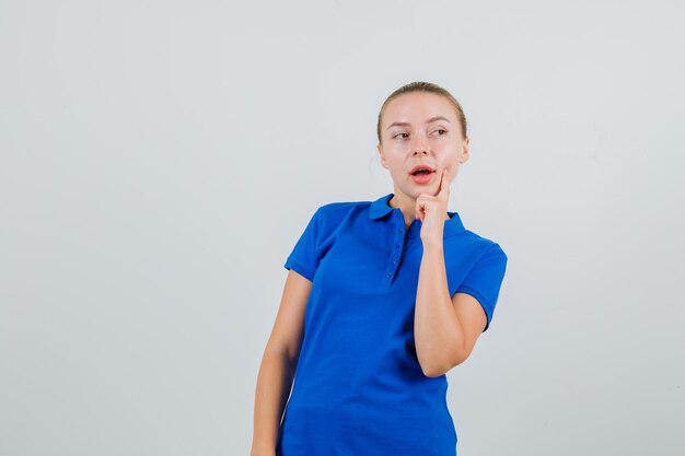 Jonge vrouw die in blauw t-shirt opzij kijkt met vinger op kuiltje en peinzend kijkt