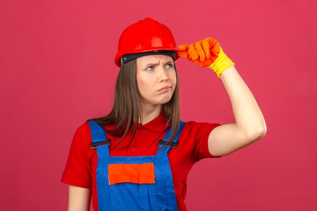 Jonge vrouw die in bouw eenvormige handschoenen en rode veiligheidshelm zijdelings met twijfelachtige en sceptische uitdrukking op donkerroze achtergrond kijken