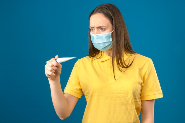 Jonge vrouw die in geel poloshirt en medisch beschermend masker thermometer in paniek op geïsoleerde blauwe achtergrond bekijken