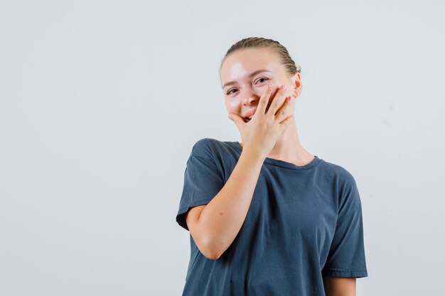 Jonge vrouw die in grijs t-shirt hand op mond houdt en vrolijk kijkt