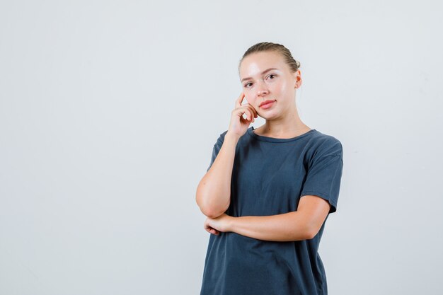 Jonge vrouw die in grijze t-shirt vingers op wang houdt en peinzend kijkt