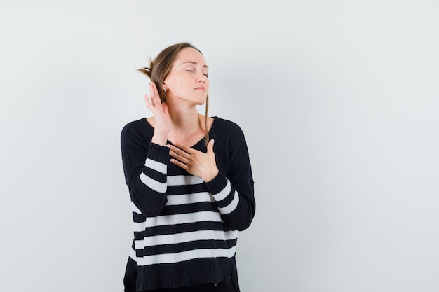 Jonge vrouw die in luisterhouding staat, een hand op de borst houdt terwijl ze de ogen sluit in gestreept breigoed en zwarte broek en er gelukkig uitziet