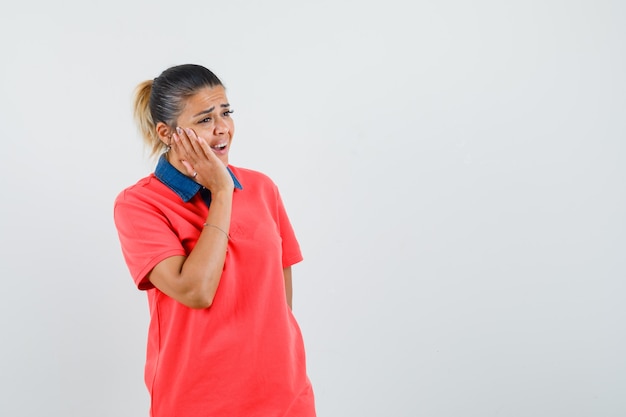 Jonge vrouw die in rode t-shirt hand op wangen houdt, kiespijn heeft en somber, vooraanzicht kijkt.