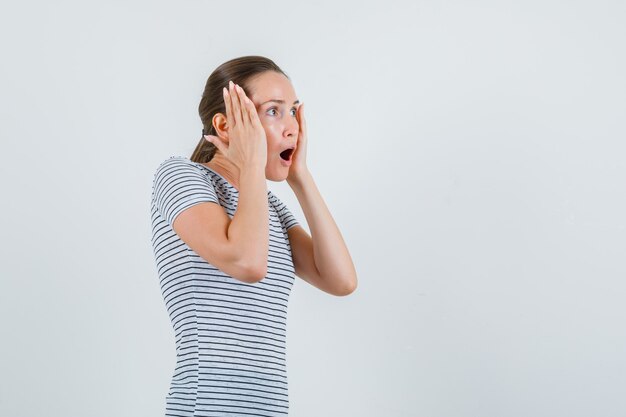 Jonge vrouw die in t-shirt de handpalmen op wangen houdt en verbaasd, vooraanzicht kijkt.