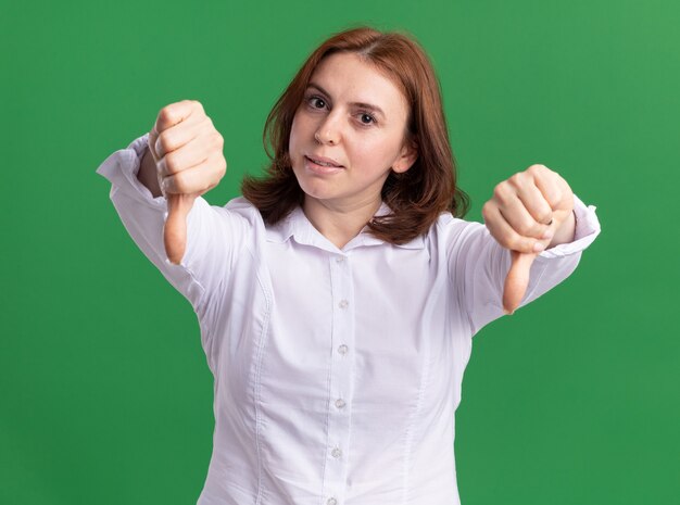 Jonge vrouw die in wit overhemd voorzijde met ernstig gezicht bekijkt dat duimen met beide handen toont die zich over groene muur bevinden