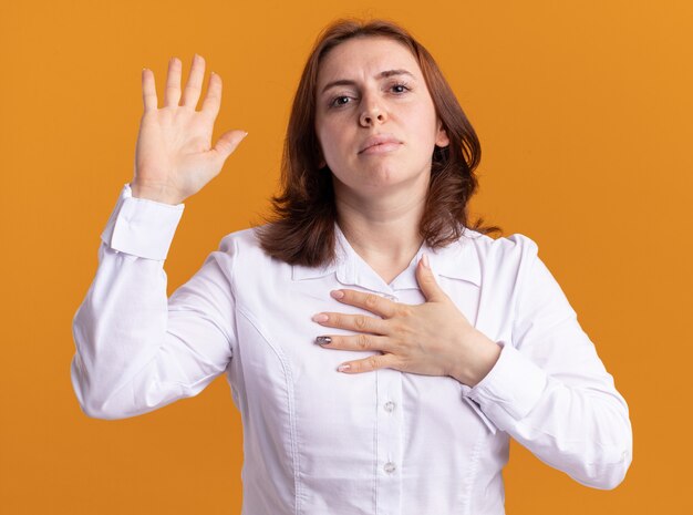 Jonge vrouw die in wit overhemd voorzijde met ernstig gezicht bekijkt die hand opheft die een belofte maken die zich over oranje muur bevindt