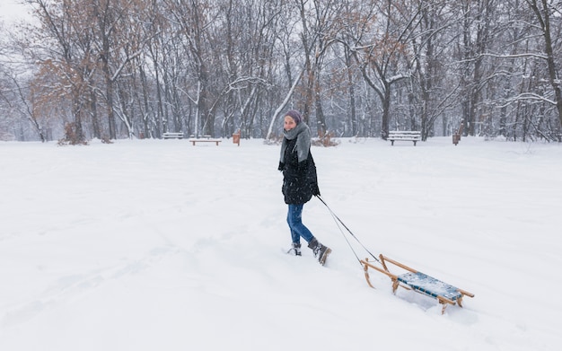 Jonge vrouw die lege houten slee op sneeuwlandschap trekt bij bos